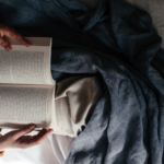arms and legs of an unseen person reading a book in bed with a blue blanket