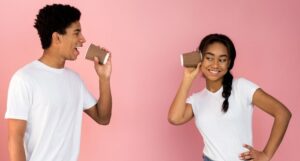 two brown-skinned teens wearing white t-shirts talking through white coffee cups with cardboard sleeves line tin can phones