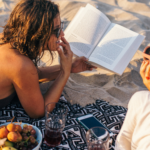 a photo of two women laying on the beach. One is reading a book