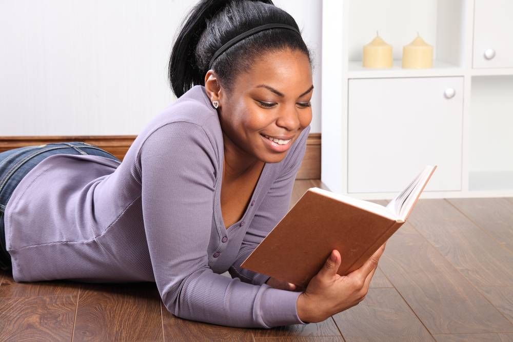 Beautiful young black girl, big smile, wearing jeans and purple top, lying on the floor at home, reading a book.