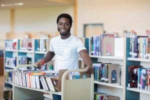 Portrait of young male librarian with trolley of books in library