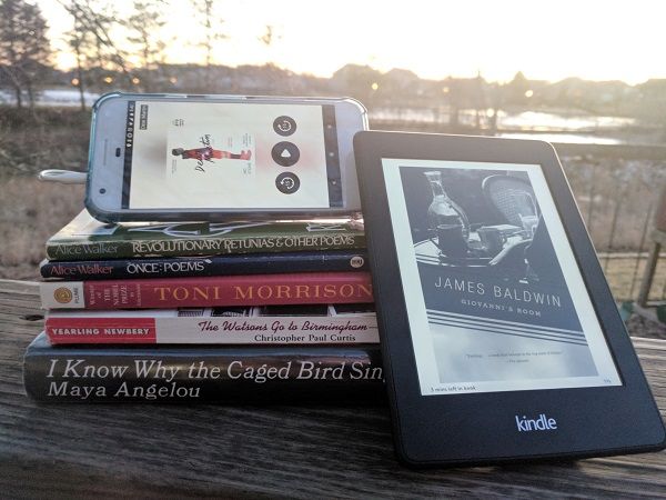 Stack of books by African-American authors listed in the article for the author's #ReadingBlackout. One is on a kindle leaning against the stack, and another is an audiobook on a phone screen set on top of the stack.