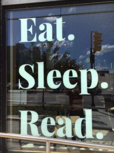 A bookstore window reflecting a city street with the words Eat. Sleep. Read. printed in big white letters.