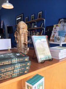 A stack of Midnight in the Garden of Good and Evil next to a stand displaying Truman Capote's In Cold Blood and a stone figurine of an elongated bald headed man