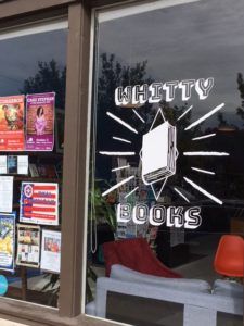 A window with Whitty Books logo and several flyers, with bookshelves and padded chairs visible through the window