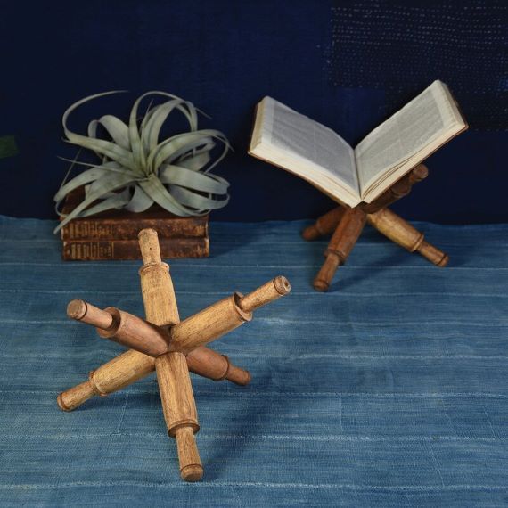 Two portable book stands shaped like jacks. A book sits on one of them. A small plant is in the background. Link: https://secure.img1-fg.wfcdn.com/im/26018860/resize-h800%5Ecompr-r85/9443/94435879/Library+Desktop+Book+Stand.jpg
