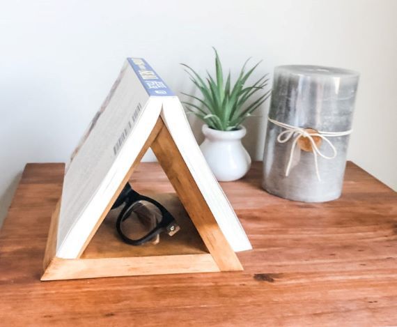 Triangular, portable book holder. A softcover book rests on top, and glasses sits inside holder. Holder, small plant, and candle sits on a table. Link: https://i.etsystatic.com/22100519/r/il/0a7c3b/2244193499/il_794xN.2244193499_h5gj.jpg