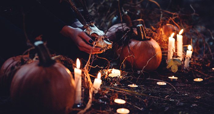 a photo of a pair of brown-skinned hands with black nail polish holding an animal skull; there are jackolanterns, lit candles, and bits of hay and leaves in the surrounding area