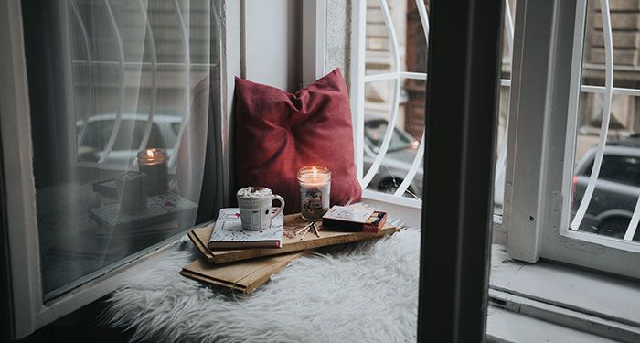 a photo of a reading nook in a window. there is a fluffy white pillow, and all the window trim is white as well. a crimson pillow sits in a corner, and there is a small book onto of a wooden rest, with a cup of hot chocolate on top and a glowing candle next to it.