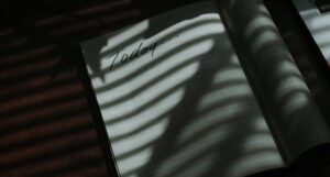 an open notebook in the shadows of window blinds. The left page of the notebook says "Today..." but is otherwise blank. https://unsplash.com/photos/6tCiSN8LX7w