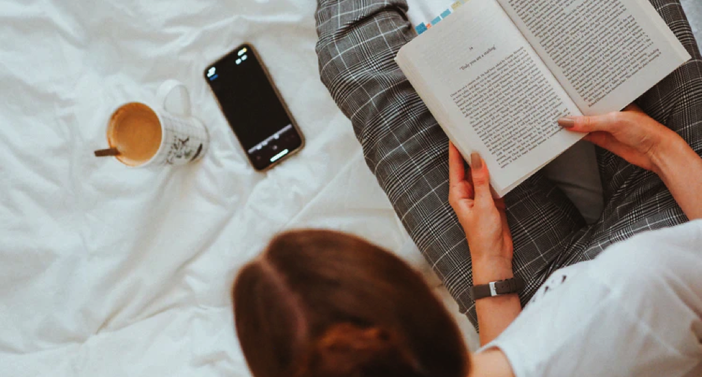 person in white shirt and plaid pants with a book in their hands as they glance at their phone https://unsplash.com/photos/X6hQVWGodOU