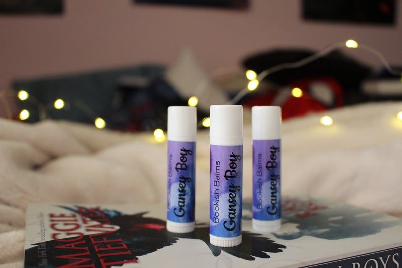 A set of three Gansey Boy labeled lip balms standing on top of The Raven Boys novel.