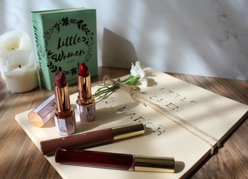 Four lipsticks on top of bound paper labeled Little Women, with the novel Little Women in the background next to two white candles