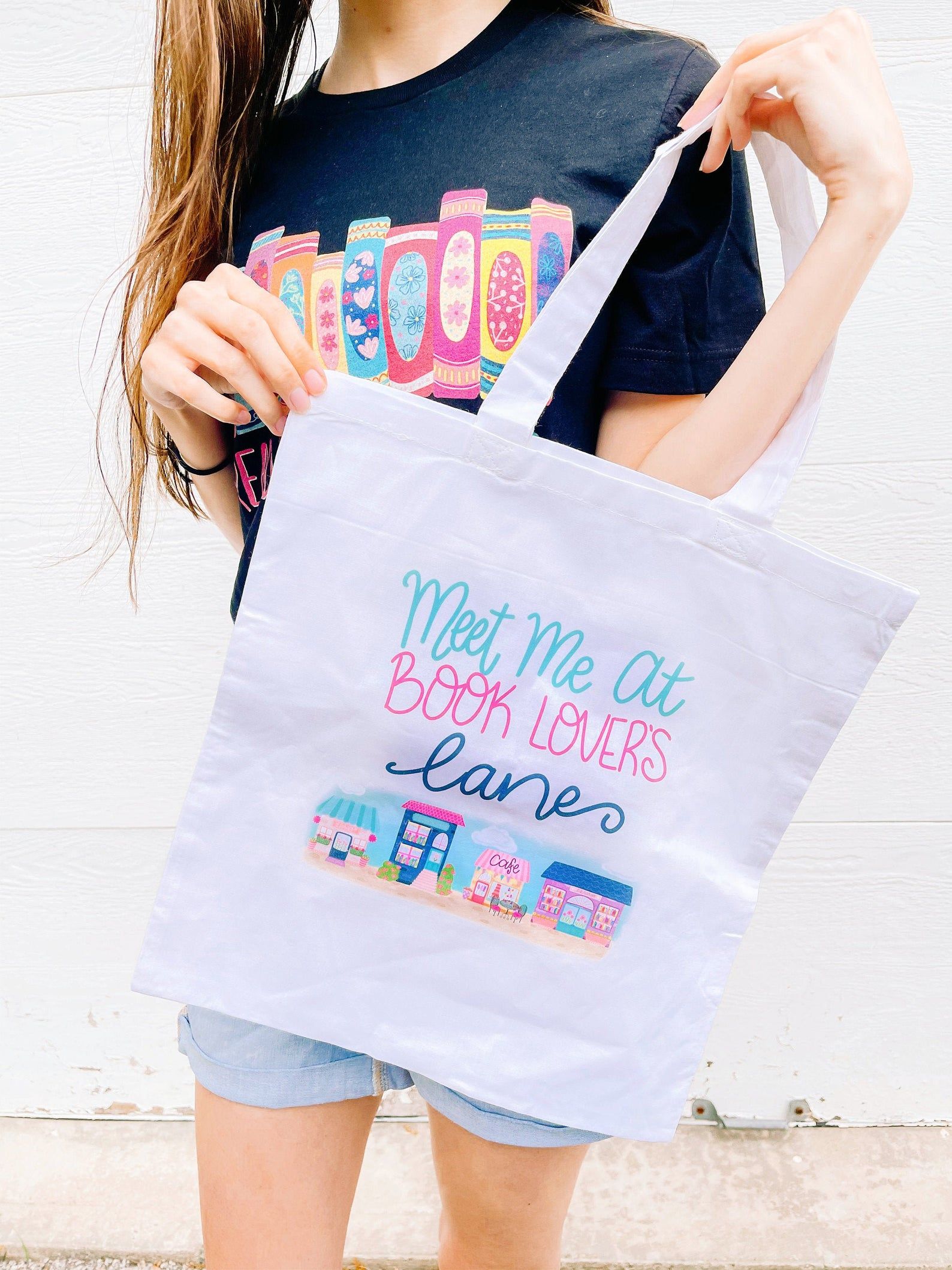 A woman holding up a tote bag. Tote bag is light purple in colour, includes a colourful image of a town and its buildings, and in colourful letter says "meet at book lovers lane" on top of the image. 