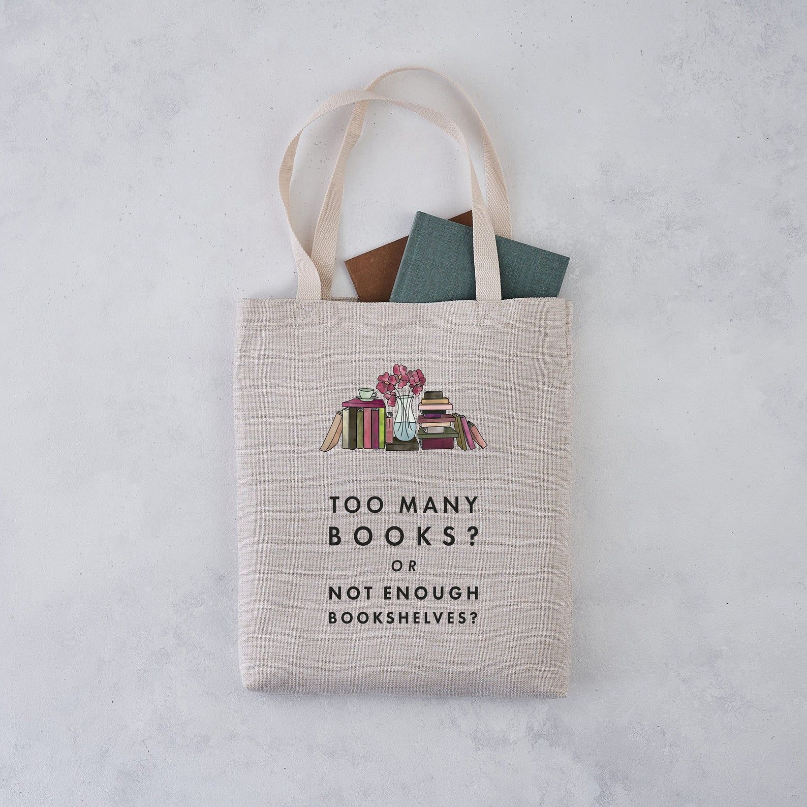 A tote bag with books peeking out rests on a gray background. The tote is light brown in colour and has an image of many books side-by-side and stacked up with a flower vase in the middle. Below, it reads: Too many books? Or not enough bookshelves? 