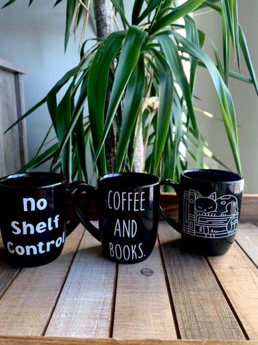 three black mugs with bookish sayings in front of a green plant