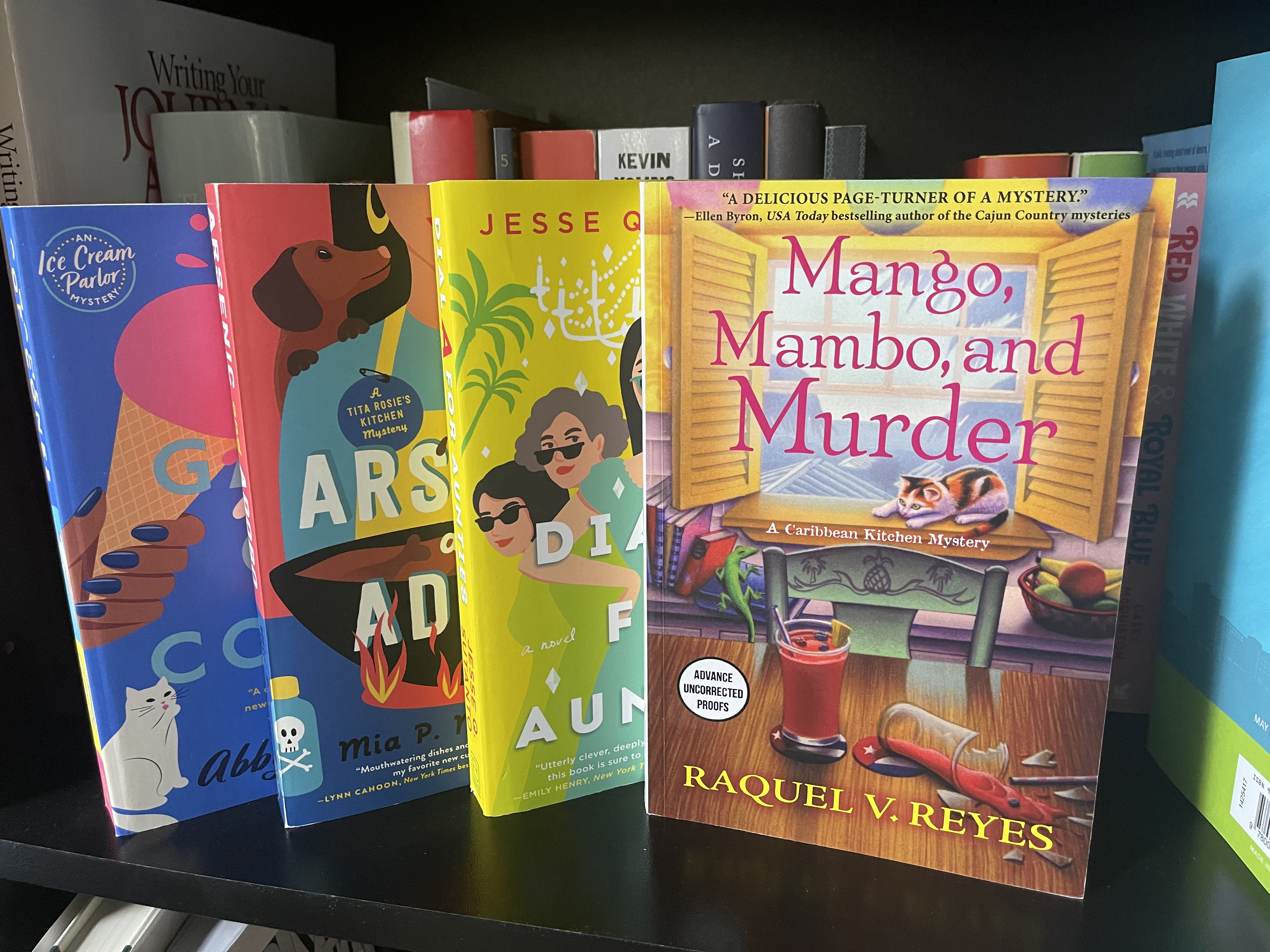 A book shelf shows the covers of four books that reflect the current trend— cozy mysteries are increasingly diverse: Game of Cones, Arsenic and Adobo, Dial A for Aunties, and Mango, Mambo, and Murder (facing forward).