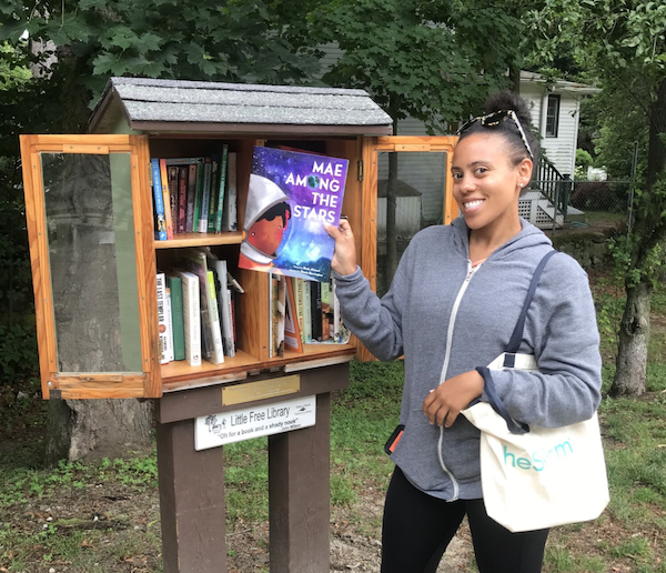 Image of a Black woman holding a book titled Mae Among The Stars in front of a Little Free Library. Image used with permission from Sarah Kamya, founder of Little Free Diverse Libraries