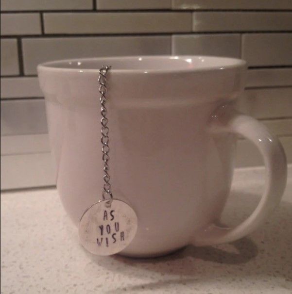 tea infuser with a silver charm at the end of a chain that says "as you wish" stamped on it