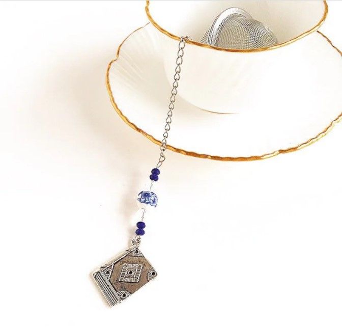 ball tea infuser with four dark blue beads, two clear beads, all split in half by a blue floral porcelain bead. At the end of the chain with the charms there is a silver book.
