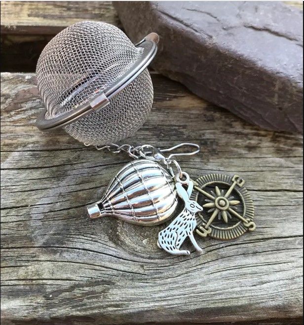ball tea infuser with three charms on it: a hot air balloon, a rabbit, and a compass rose