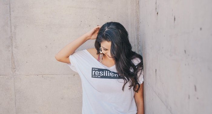Image of a woman with brown skin against a cement wall, with a shirt reading "resilient"
