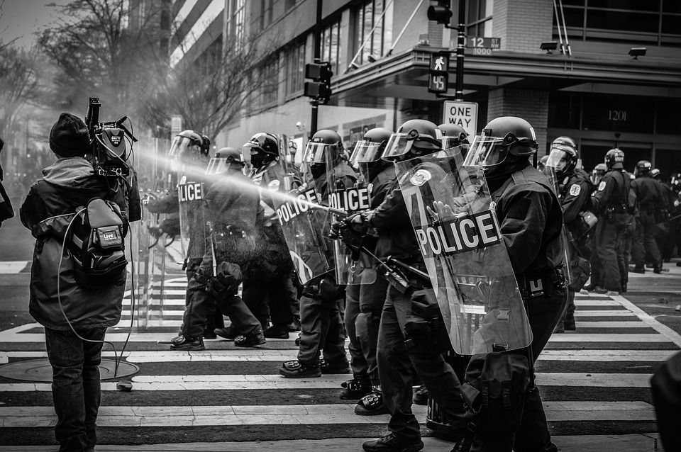 Black and white image of a line of police in riot gear, carrying shields, walking in a road in a city. One of the police officers is spraying pepper spray towards a person with a backpack and video camera