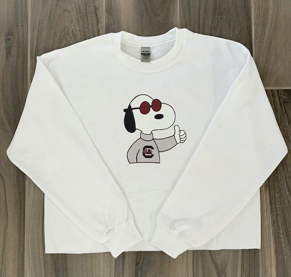 A white sweatshirt featuring Snoopy. Snoopy is in his Joe Cool persona, wearing a University of South Carolina sweatshirt and giving a thumbs up.