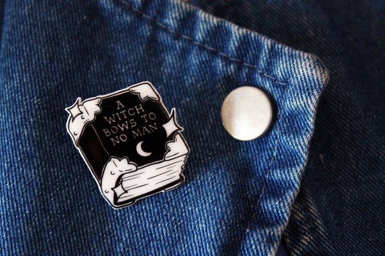 Enamel pin in the shape of a book reading "A witch bows to no man."
