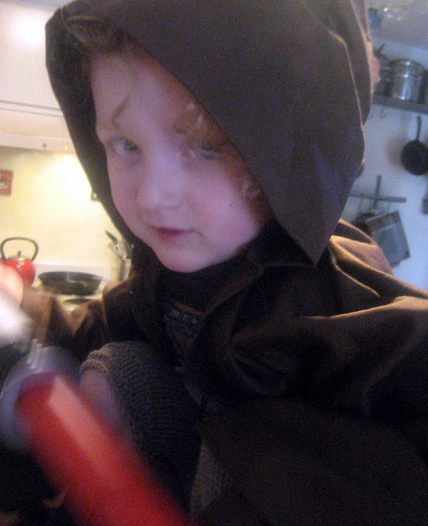 a photo of a child wearing a brown hooded robe and carrying a red plastic lightsaber
