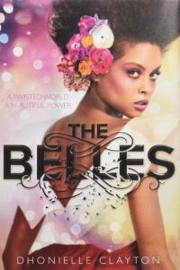Book cover for The Belles, showing a pink and yellow blurred background, a black woman with afro hair and a floral fascinator attached to her hairstyle. She wears a white off-the-shoulder top. The title is superimposed over her.