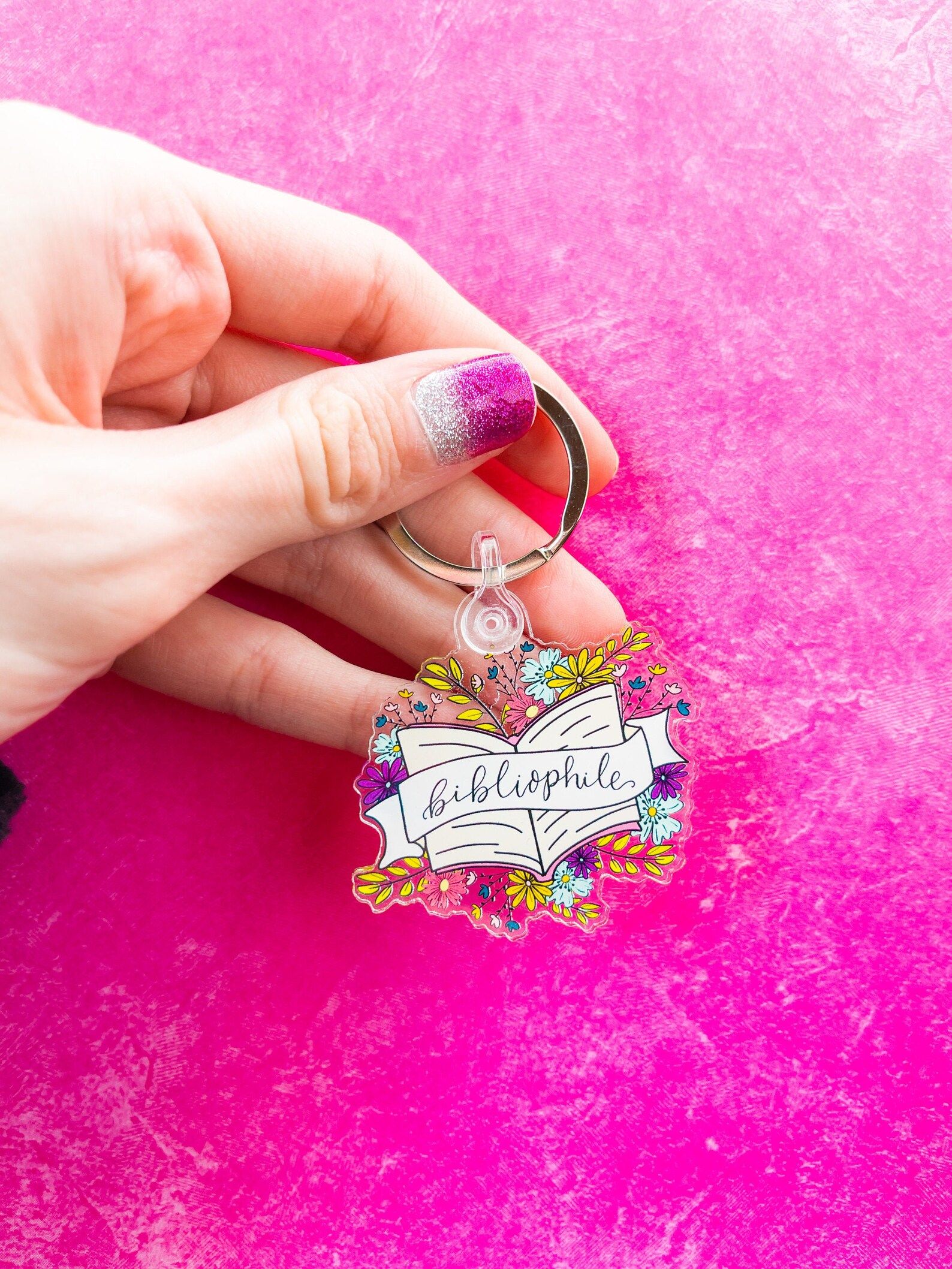an acrylic keychain depicting flowers, an open book, and a banner that reads "bibliophile"