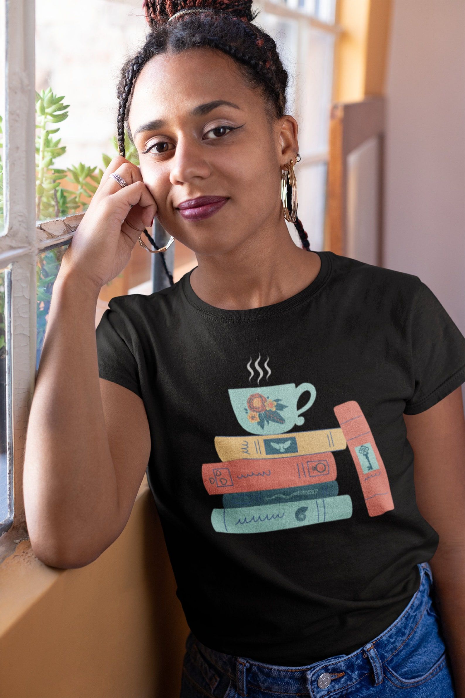 Image of a Black woman wearing a black t-shirt. The shirt has a green tea cup on top of four books in yellow, orange, and green. There is a fifth book leaning against the stack. 