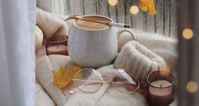 A photo of coffee, glasses, and a cozy sweater by an open book. Rain is visible through the window and leaves are scattered around