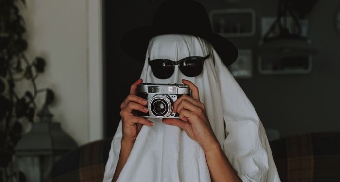 person dressed like a ghost with a sheet over their head plus sunglasses and a hat. The person is holding an old fashioned camera