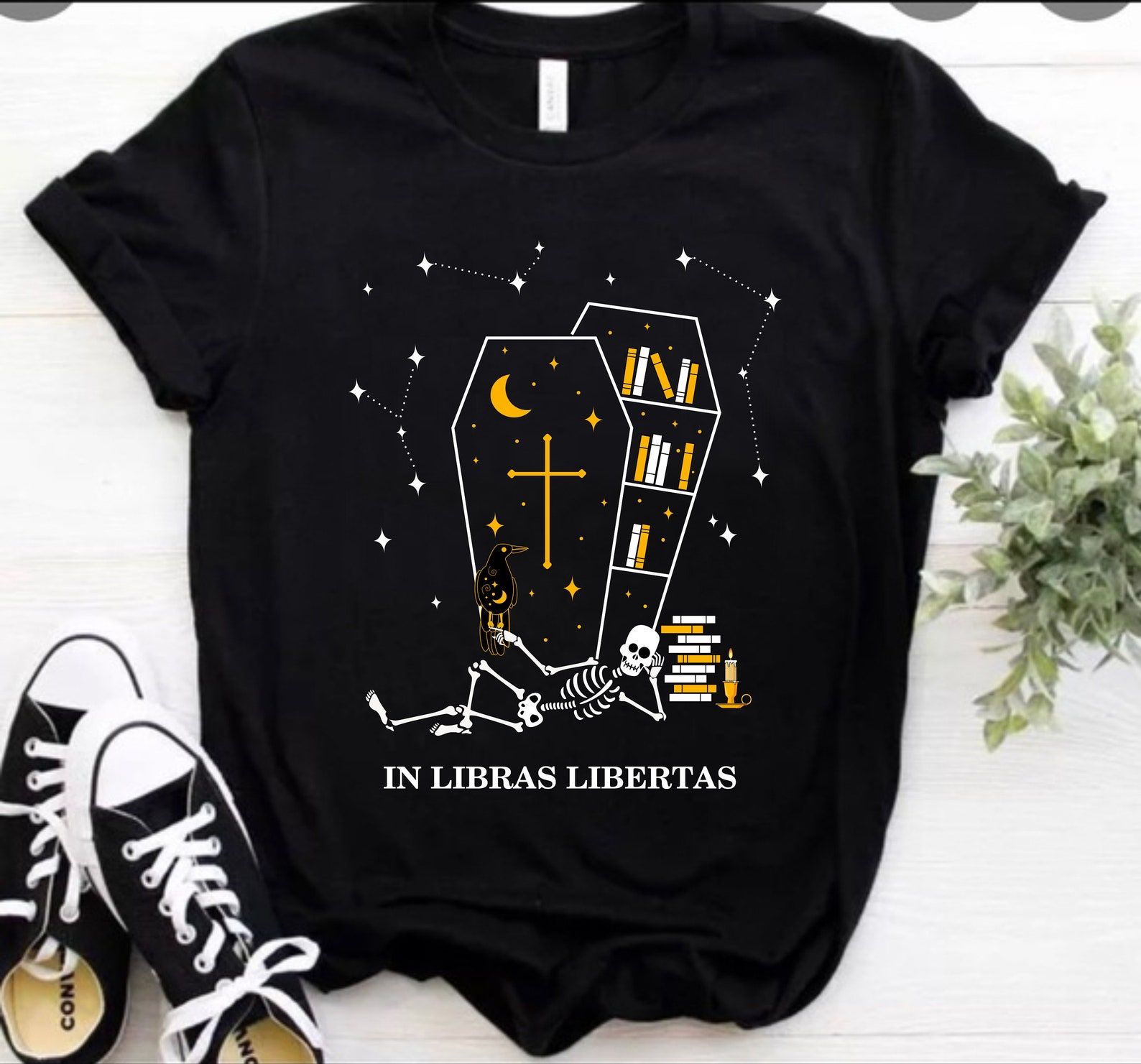 Image of a black t-shirt. It has a coffin filled with books on it, and a skeleton lying in front of that. It reads "in libras libertas." 