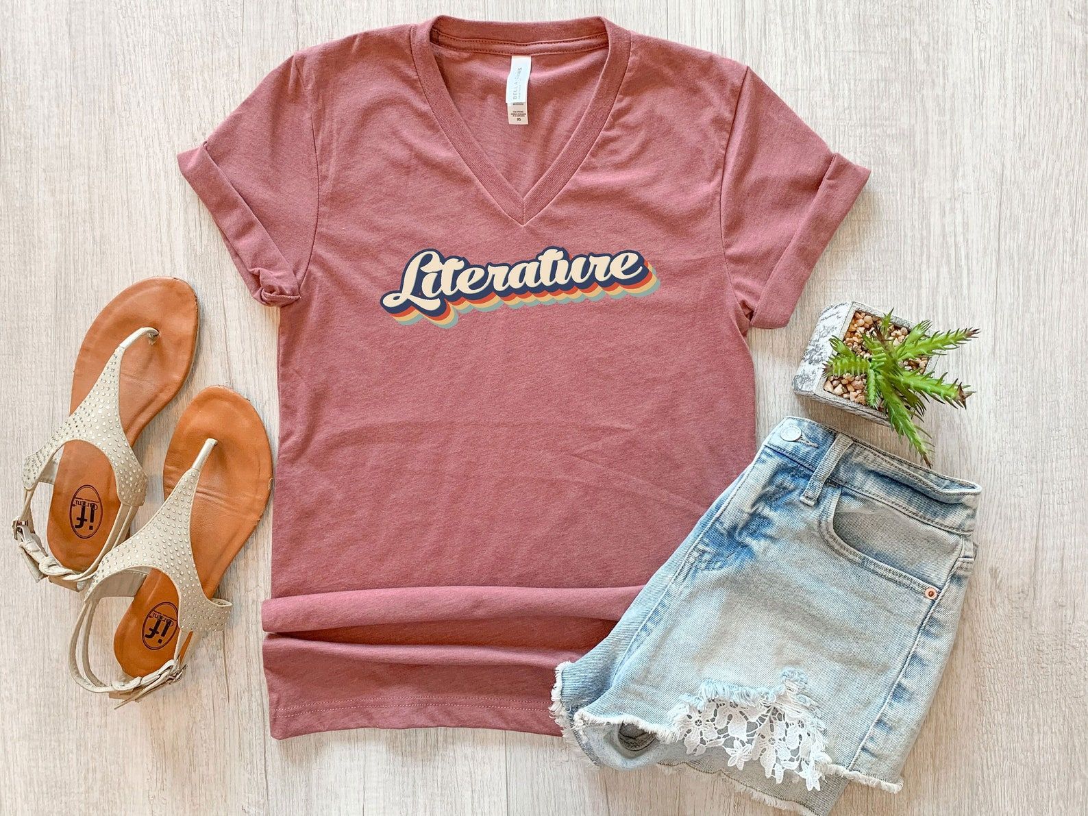 Image of a pink v-neck shirt with the word "literature" on it. The letters are shadowed in rainbow colors. 