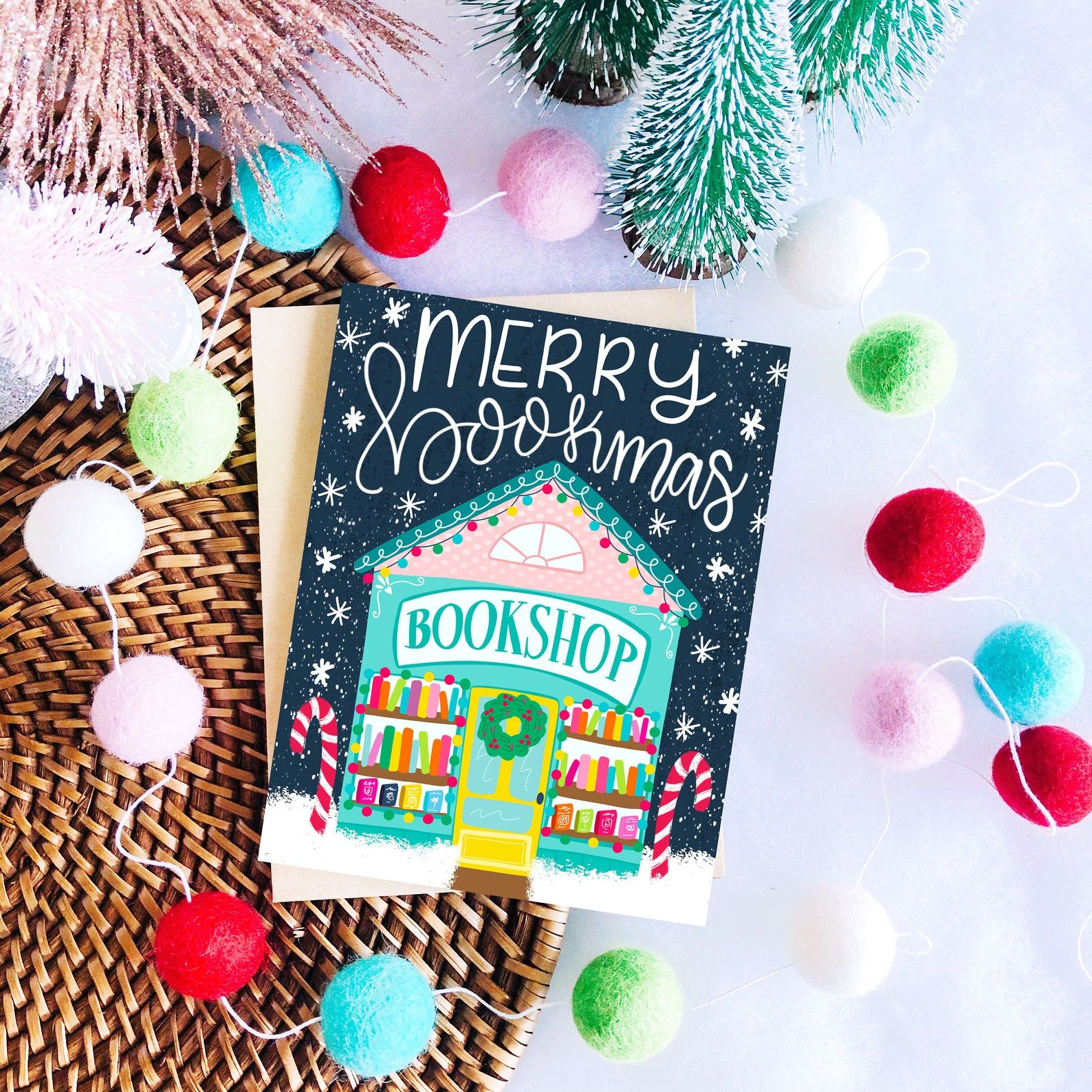 A dark blue card that reads "Merry Bookmas" and depicts a teal bookshop with colorful books pines and pink, red, and green accents.