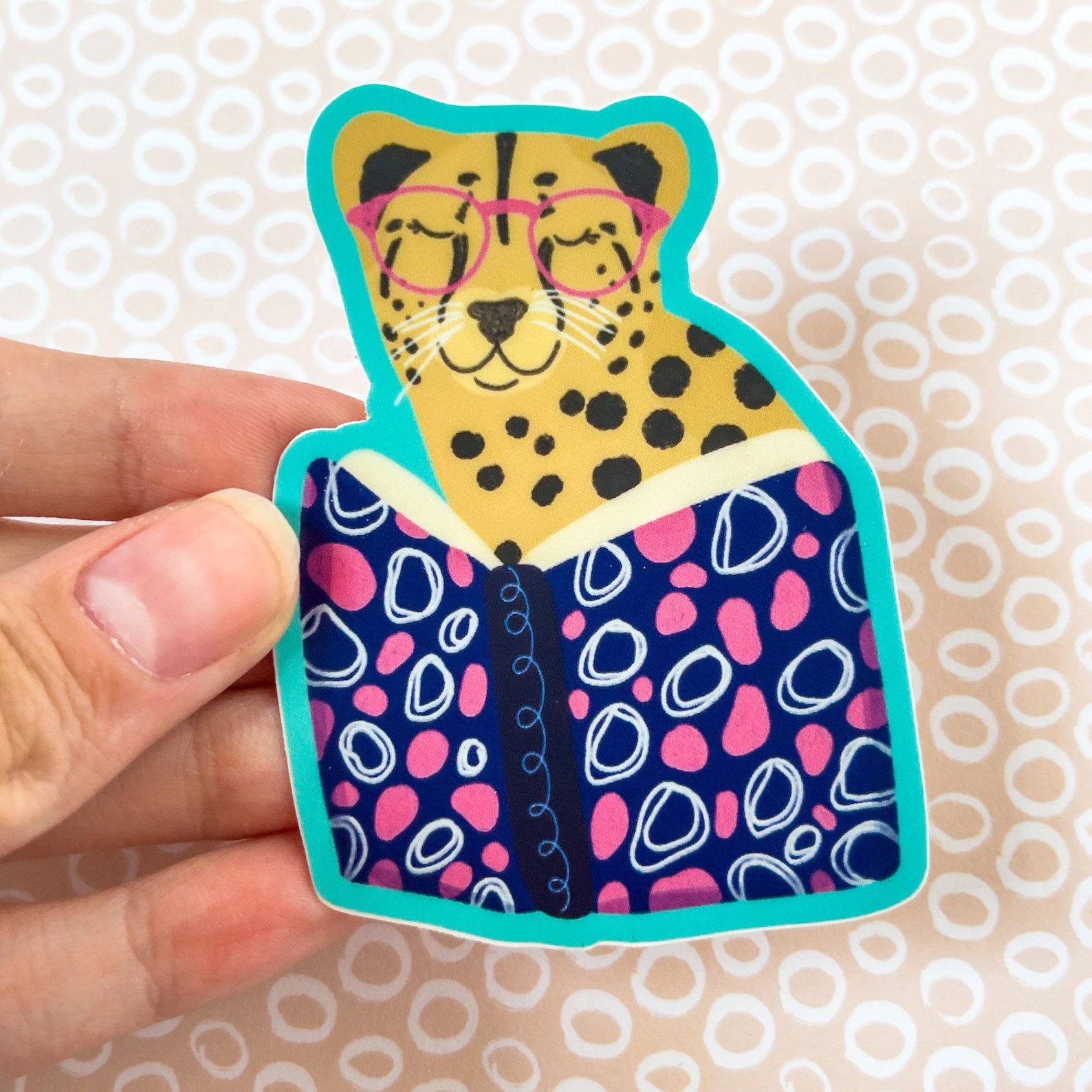 A sticker depicting a cheetah in pink glasses reading a book that is decorated with blue, pink, and white squiggles.