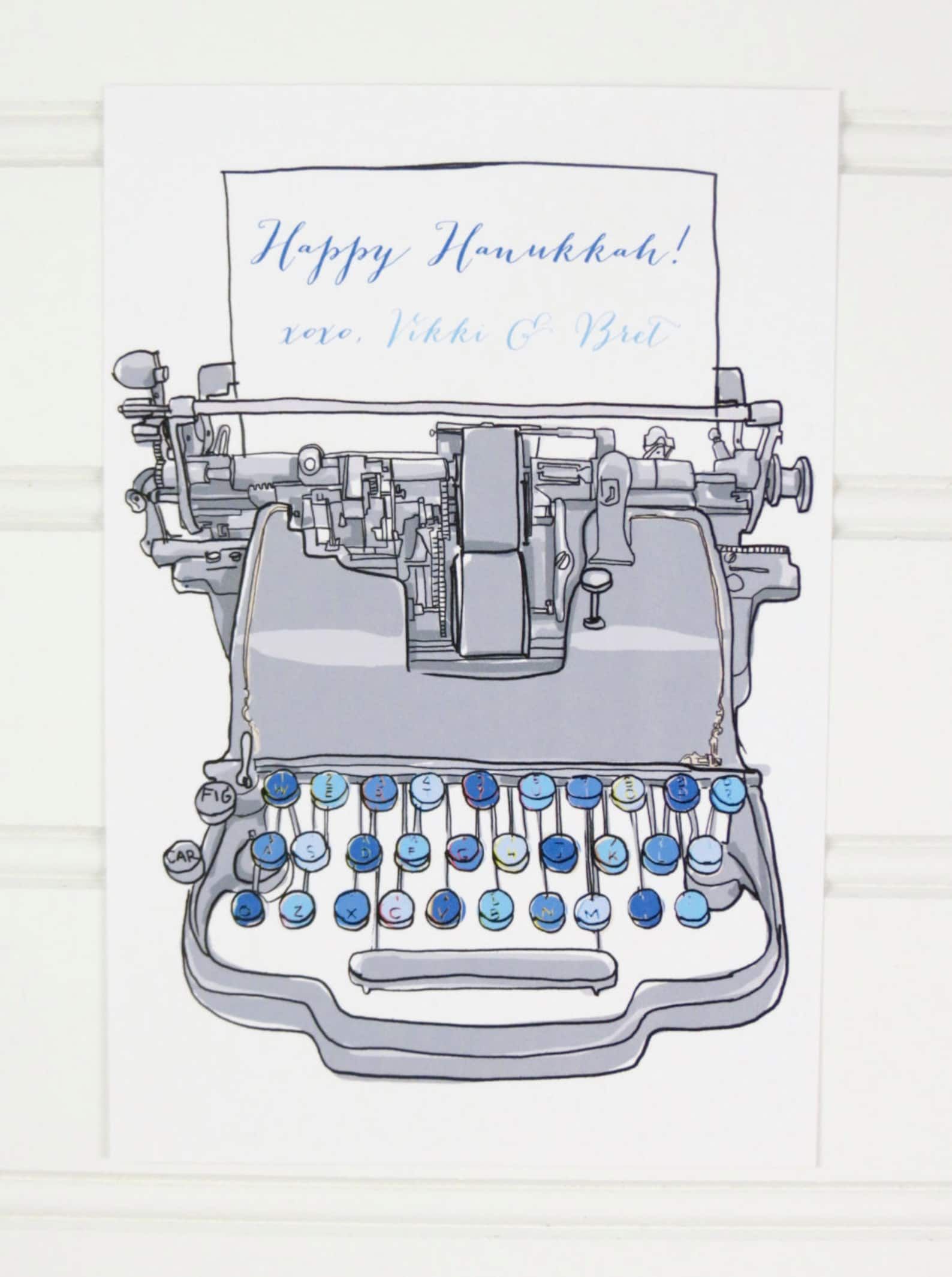 A vintage blue typewriter typing out "Happy Hanukkah" with space for a personalized message on a white postcard