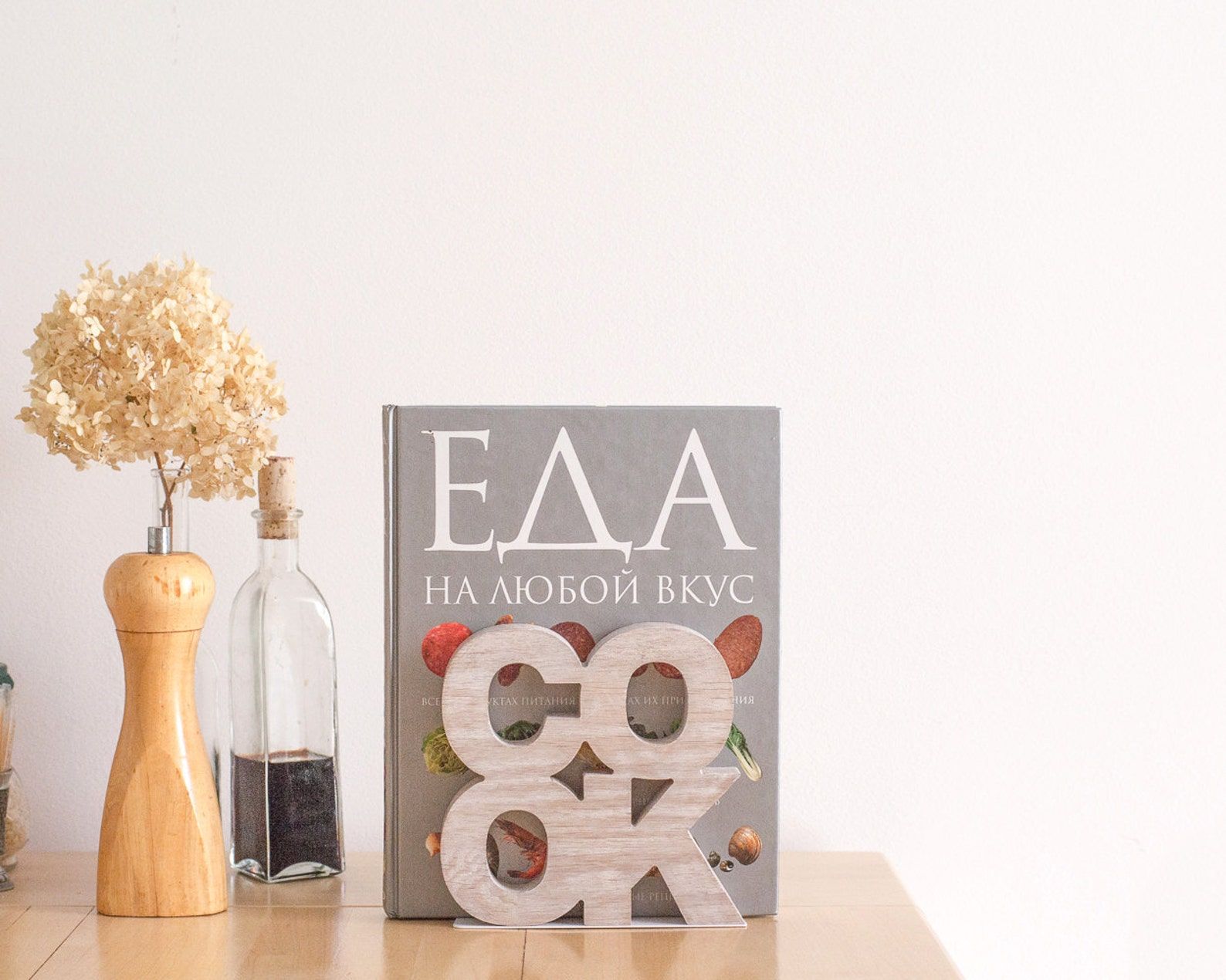 A wooden bookend with large letters that spell "Cook" laid out in a square. It's holding up a cookbook and there is a pepper grinder next to it.