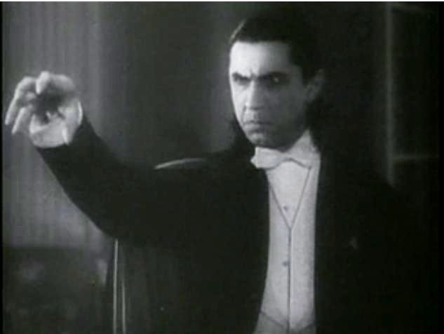 A black and white image of Bela Lugosi as Dracula in the 1931 movie of the same name. He wears a white tie tuxedo and a dark cape, extending his hand in a creepy claw. 