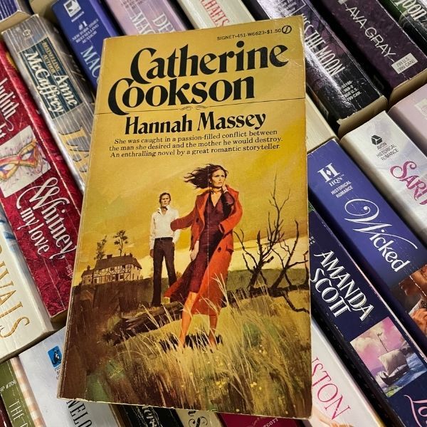 the cover of a book called Hannah Massey, with a woman running from a manor in a gloomy landscape with a red coat draped loosely over one shoulder