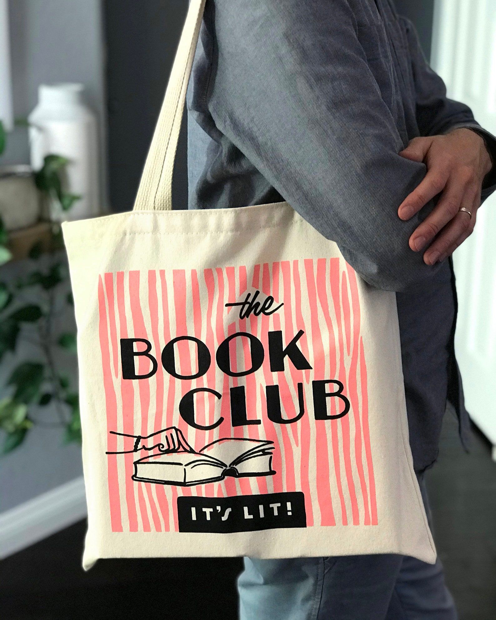 Image of a canvas tote bag. It's got pink stripes and the words "The Book Club: It's Lit" in black font, along with an image of a finger pointing in an open book. 