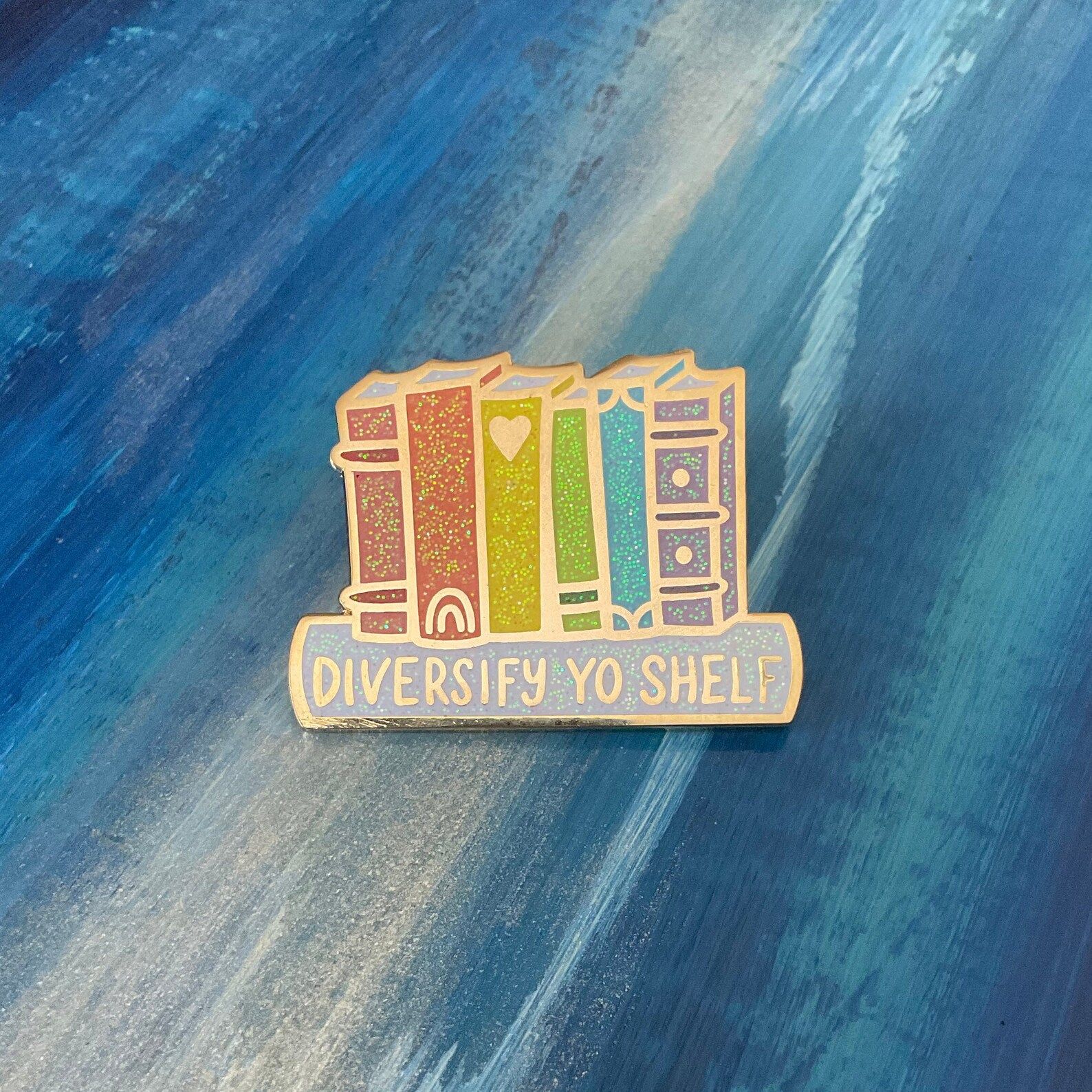 An enamel pin of rainbow book spines with the words "diversify yo shelf."