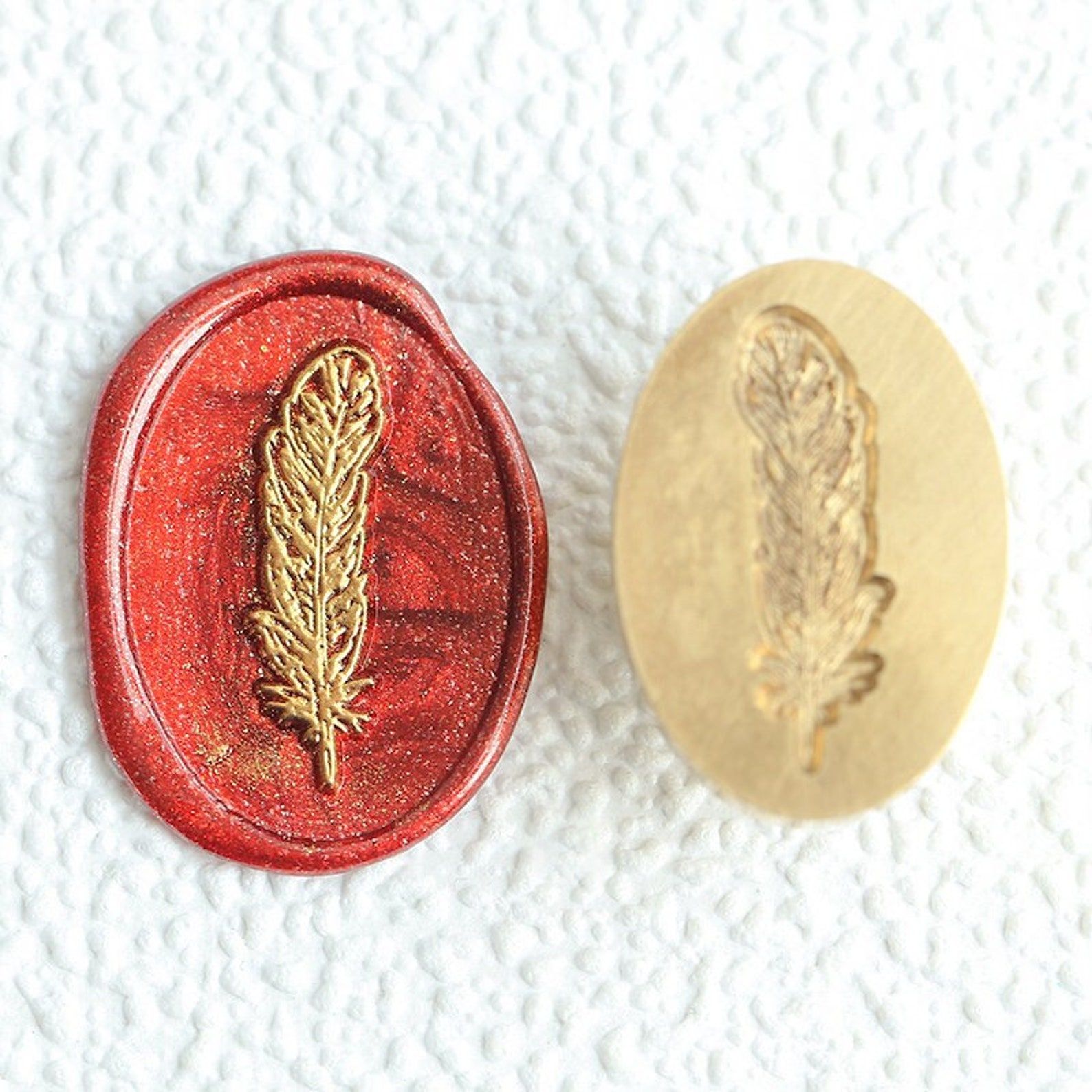 Image of a wax stamper, alongside its wax seal. The seal is red, with a gold feather in the center. 