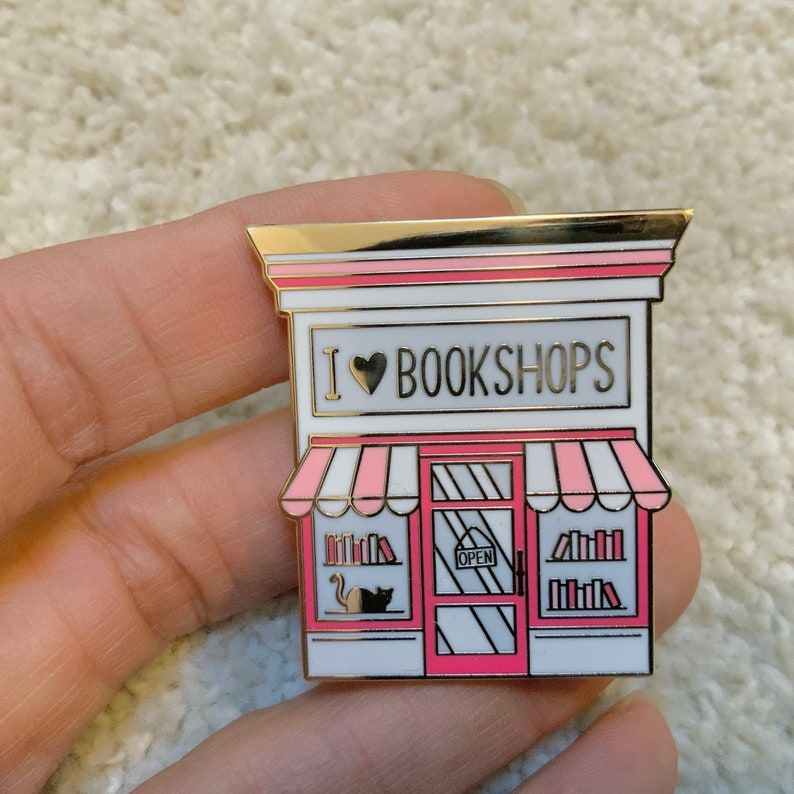 Image of an enamel pin. It's a pink bookshop store front, with the words "I <3 Bookshops" in gold. 