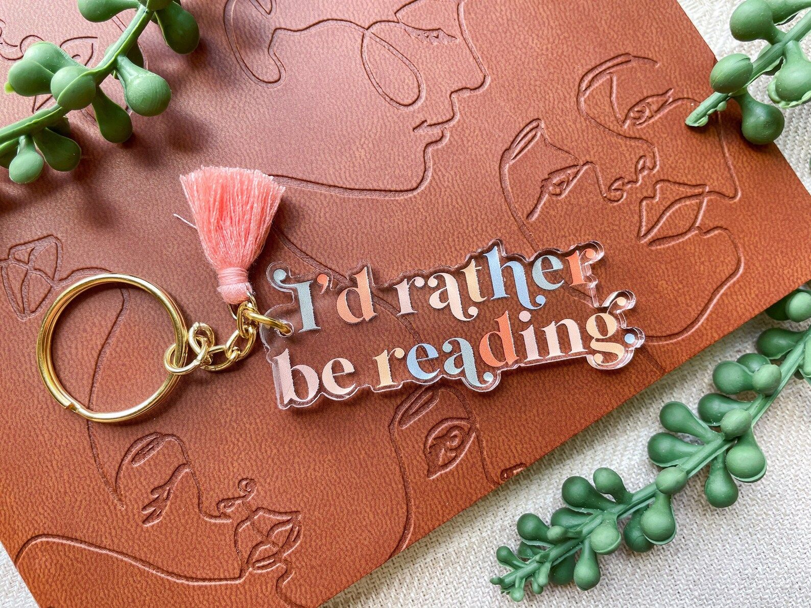 A clear acrylic keychain with pink and blue lettering that reads "I'd rather be reading" with a pink tassels on a gold keyring.