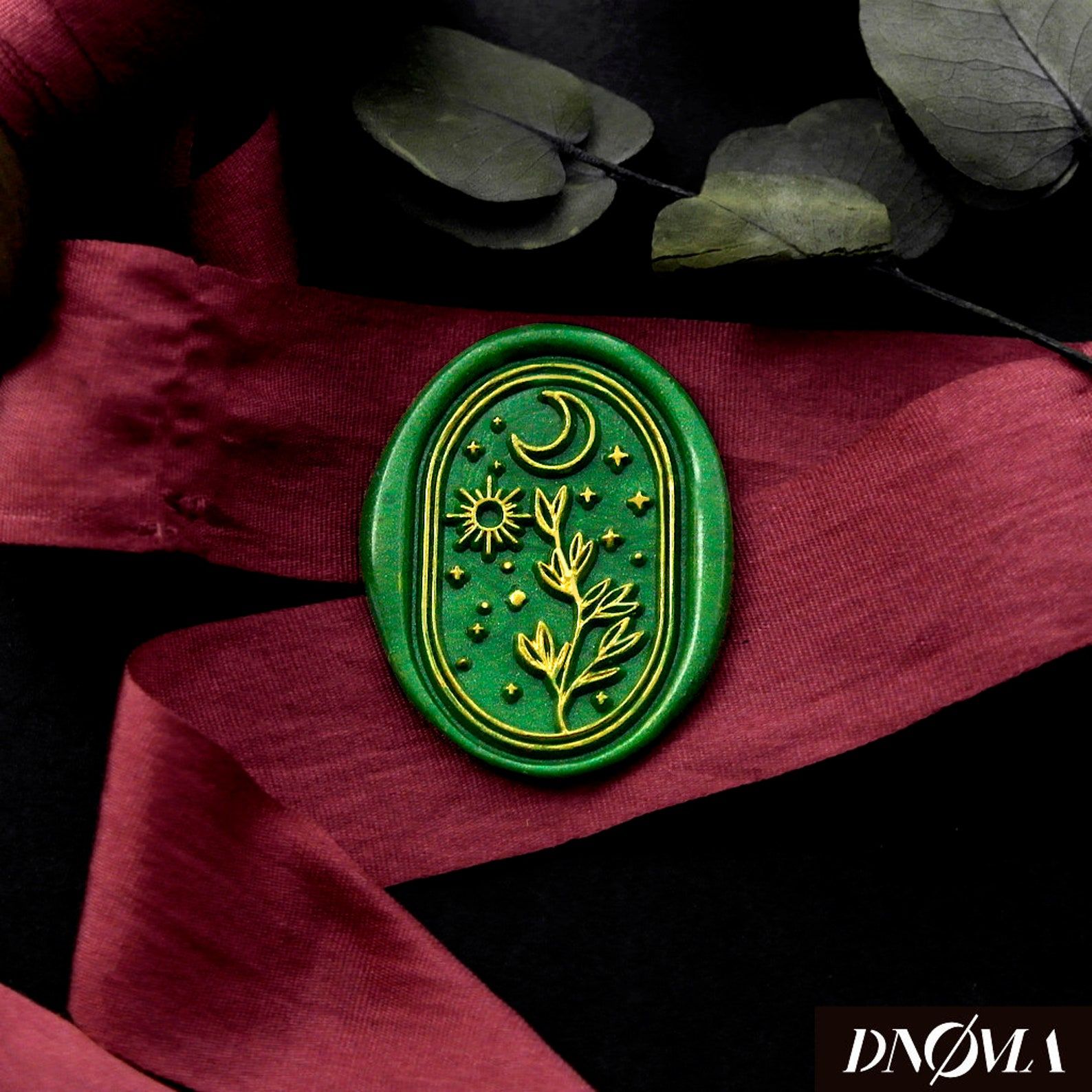 Image of a dark green wax seal, featuring a gold moon, sun, and plant. Those are in gold, and the stamp is on top of a red ribbon.  