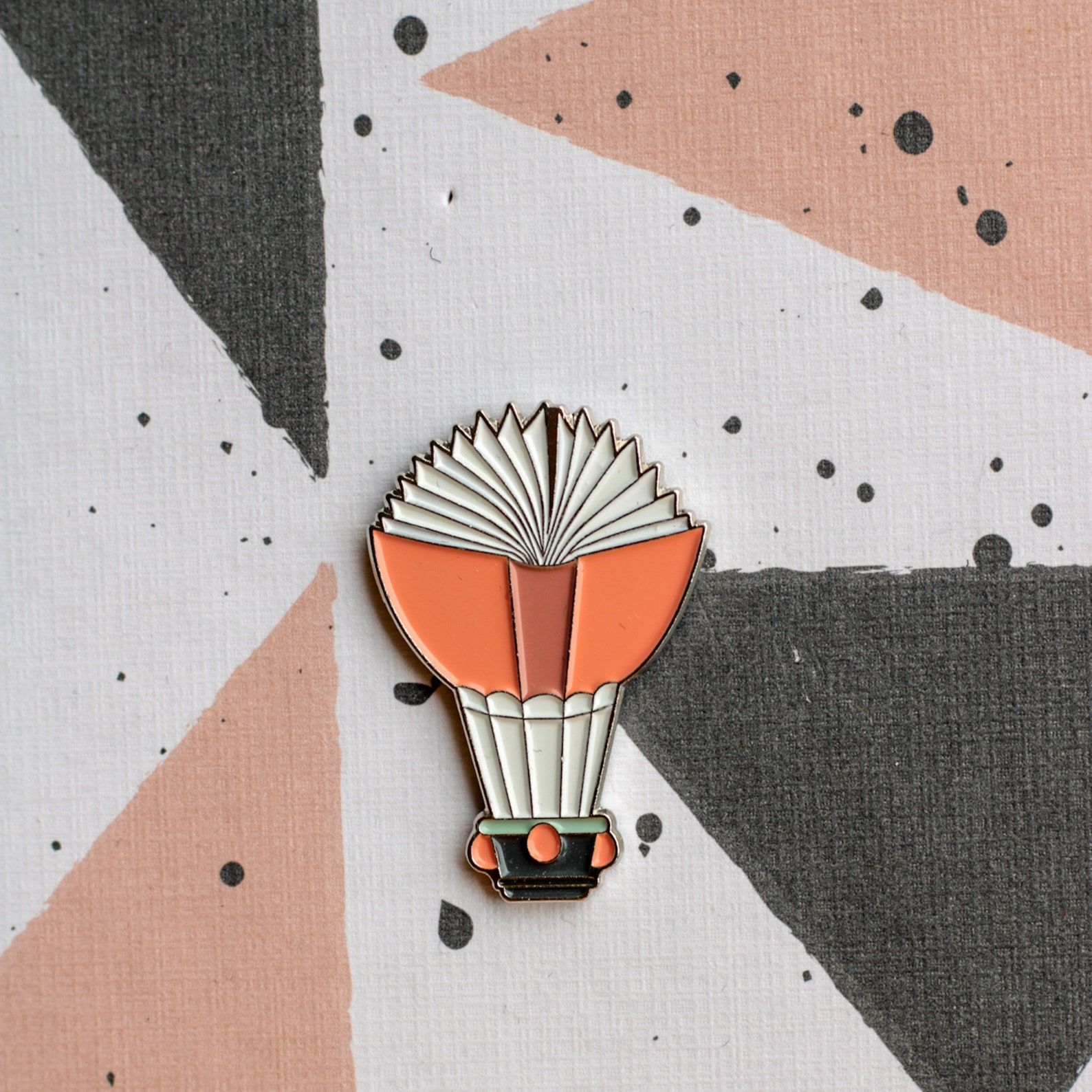 Image of an enamel pin -- it has a pink book open like a hot air balloon. 